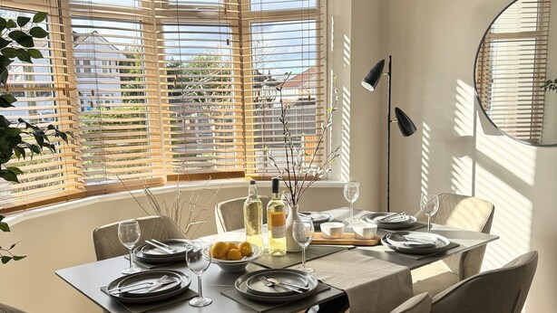 Un comedor con mesa dispuesta para cuatro personas, ventanas amplias con persianas y una lámpara colgante moderna.
