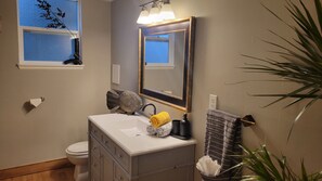 Ein Badezimmer mit einem weißen Waschbecken, einem Spiegel, einem Handtuchhalter, einer Toilette und einem Fenster mit Blick auf den Himmel.