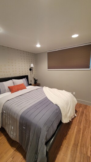 Ein Schlafzimmer mit Bett, Nachttisch, Lampe, einem Fenster mit Jalousien und einem Wandbild.