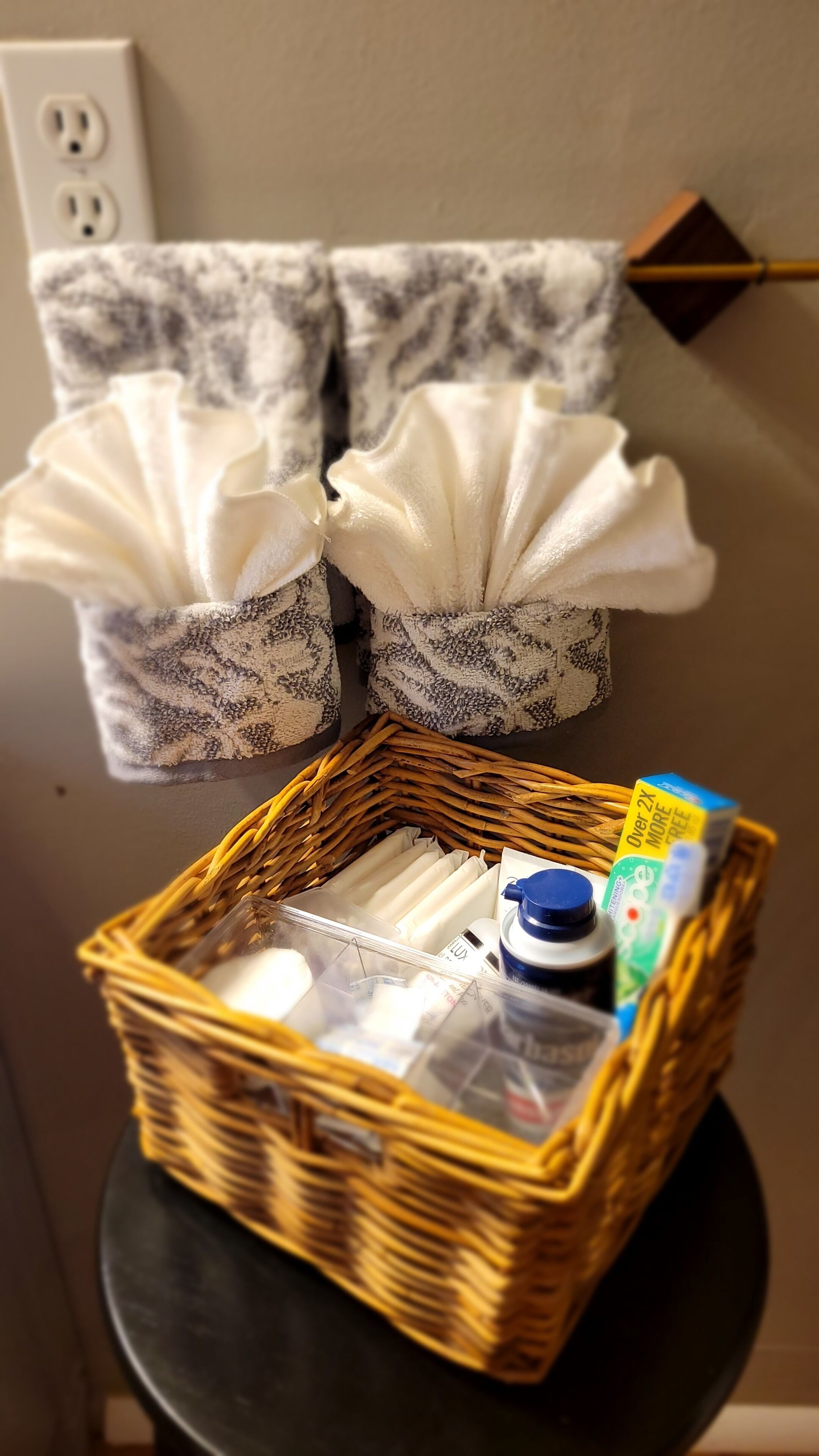 A wicker basket with toiletries on a small table, two patterned towels hanging on a wall, and a power outlet in the background.