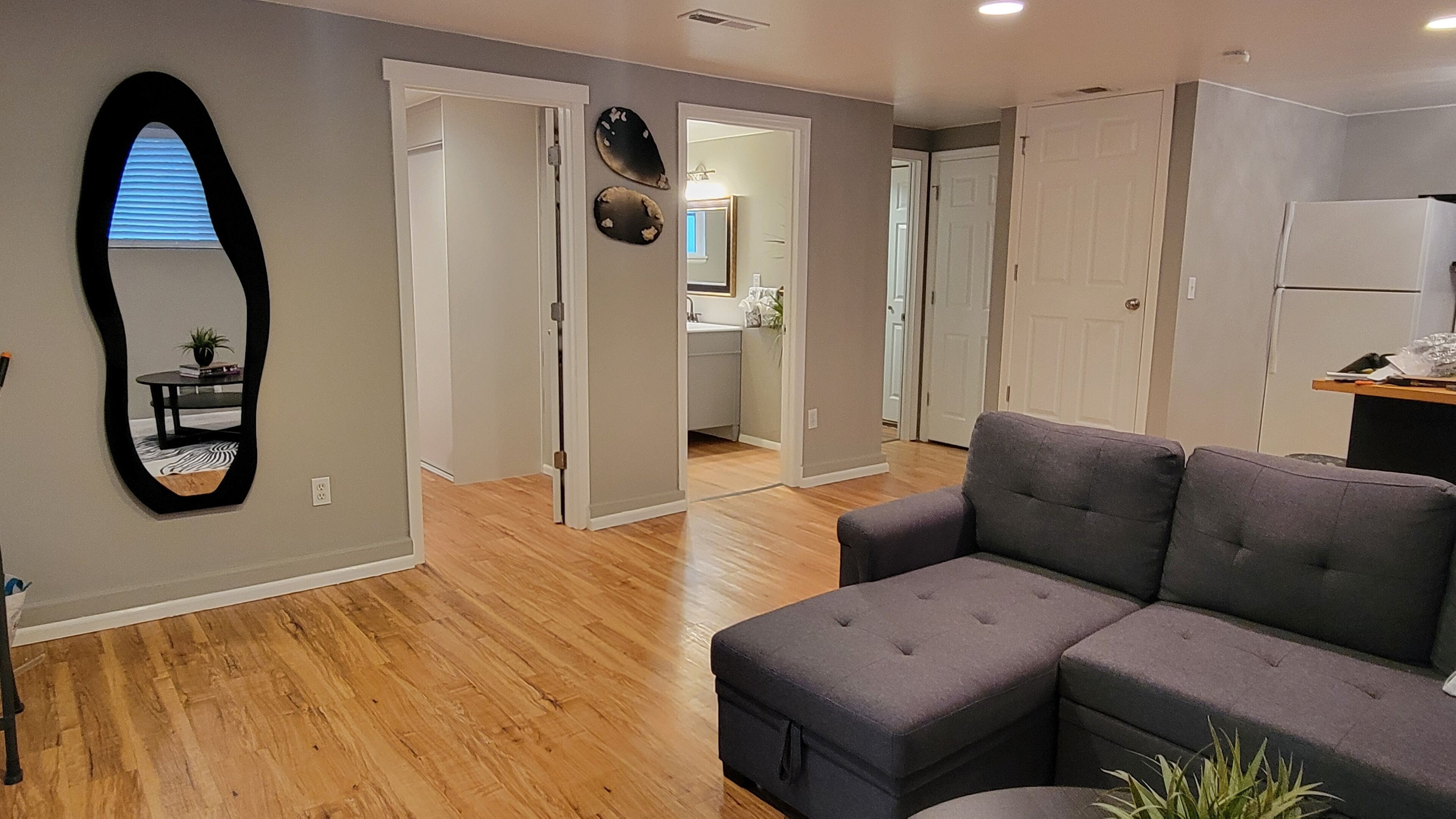A living room with a grey sectional sofa, wooden flooring, a mirror, and a hallway leading to a kitchen.