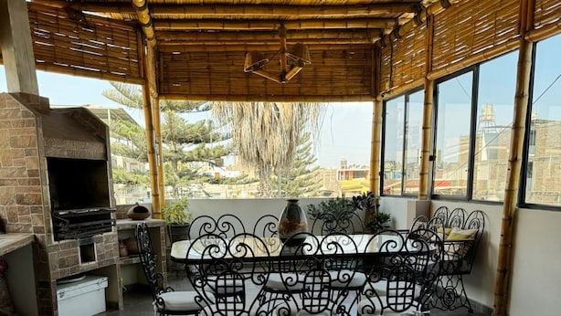 Une terrasse extérieure dotée d’un four en briques, d’une table à manger avec des chaises décorées et de grandes fenêtres offrant une vue sur les environs.