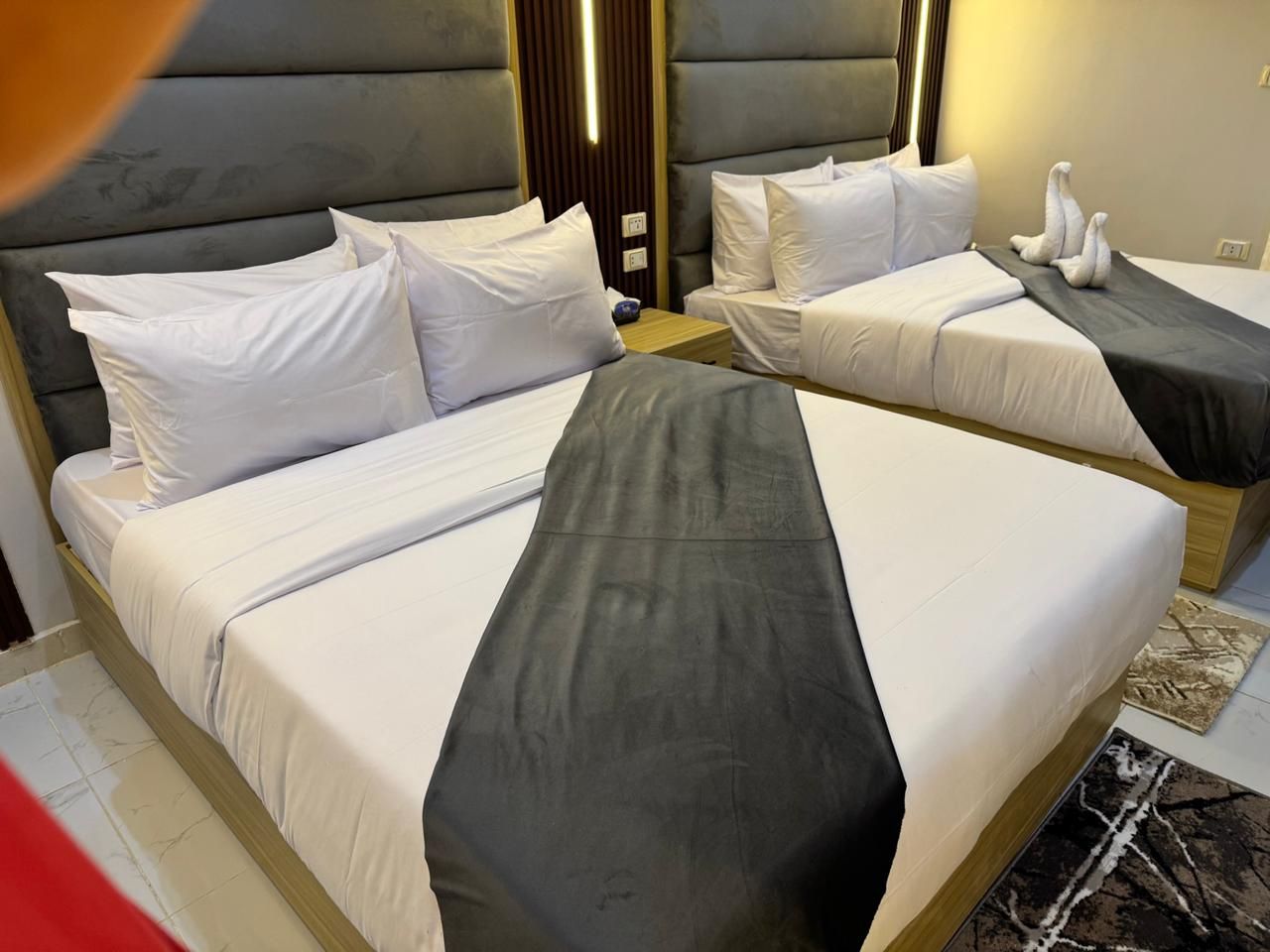 A hotel room with two beds, each with white bedding and a grey runner. There are wooden headboards and a small wooden nightstand between the beds.