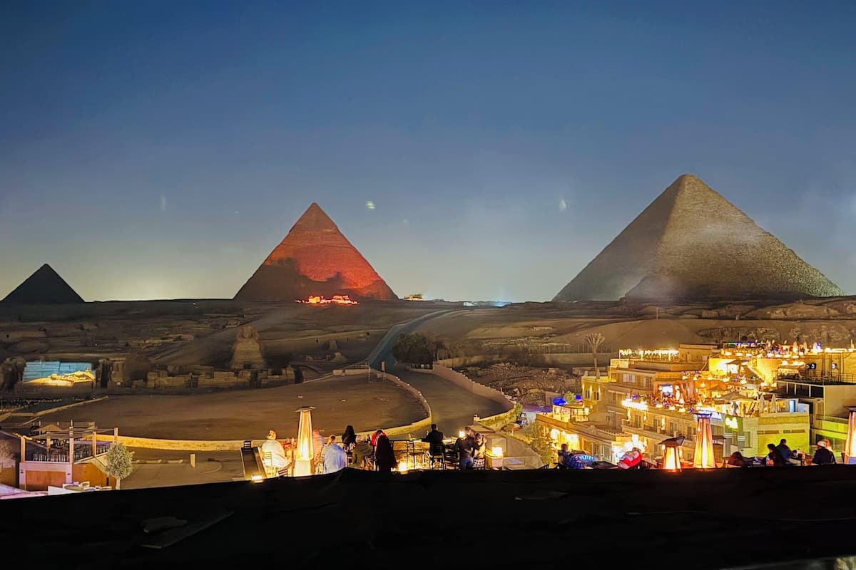 A view of the pyramids at Giza with a road leading towards them, and a well-lit area with people and structures in the foreground.