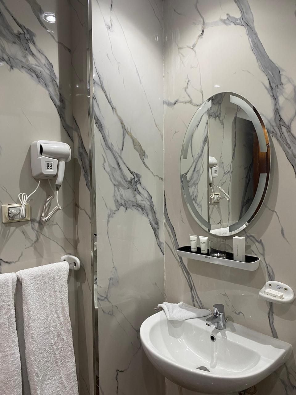 A bathroom with a marble wall, a round mirror, a white sink, a towel rack, and a hairdryer.