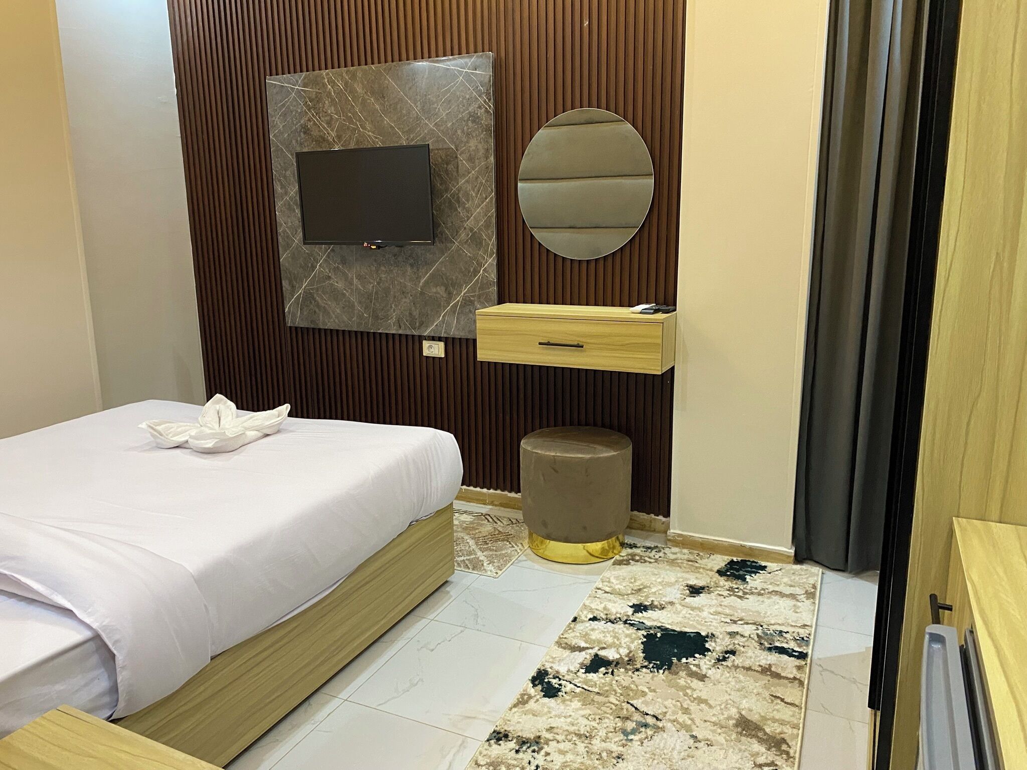 A modern bedroom with a bed, a vanity with a mirror, a small stool, and a television mounted on a wall with a marble-like texture.