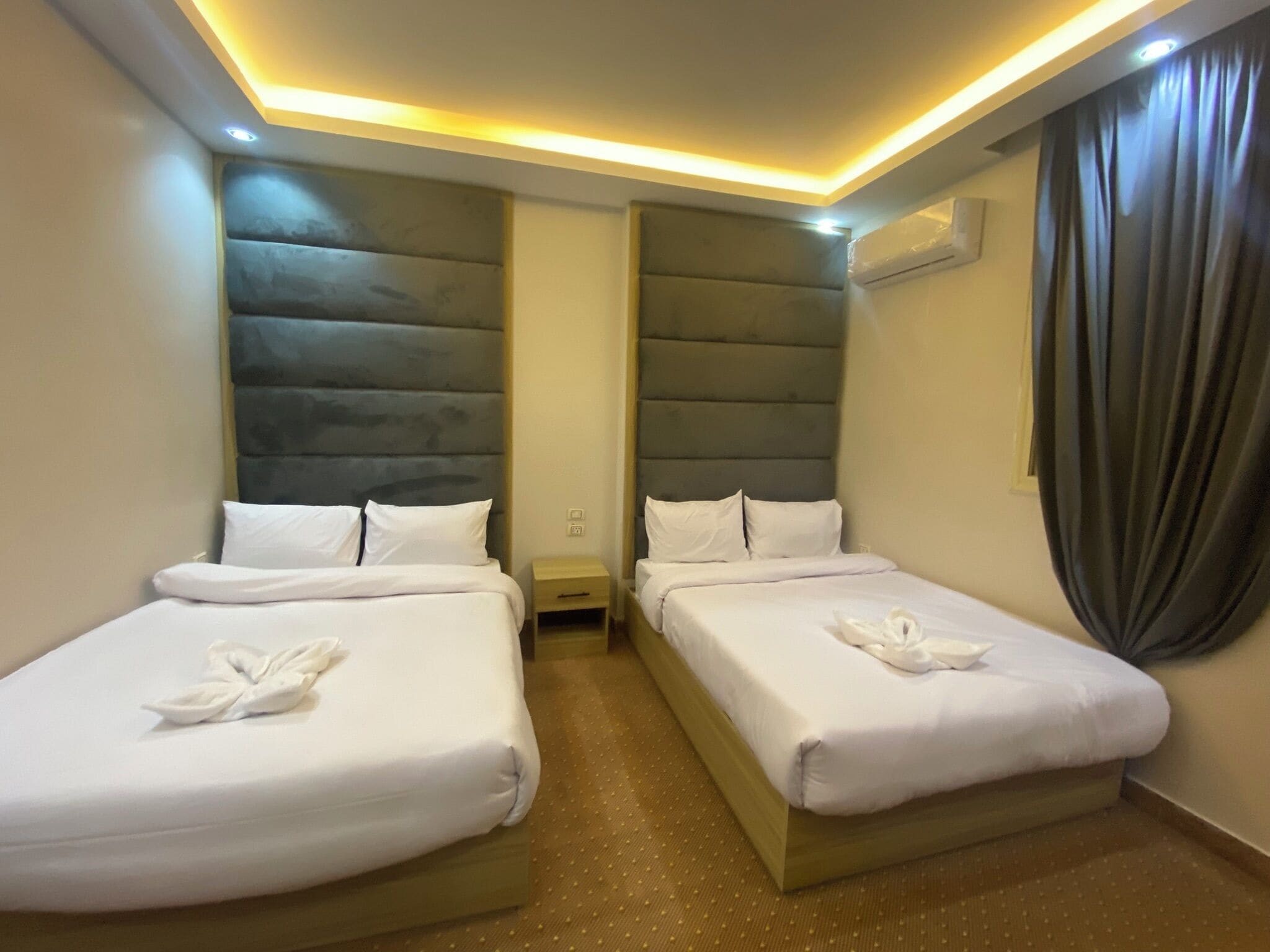 Habitación de hotel con dos camas, cada una con ropa de cama y toallas blancas, una mesita de noche pequeña y un cabecero con diseño acolchado.