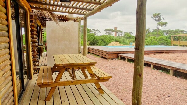 A wooden deck with a picnic table, surrounded by a wooden fence and a view of a pool and greenery. - Amazing 2-bedroom bungalow with WiFi, AC in wonderful Punta del Diablo,Uruguay
 (Punta del Diablo)