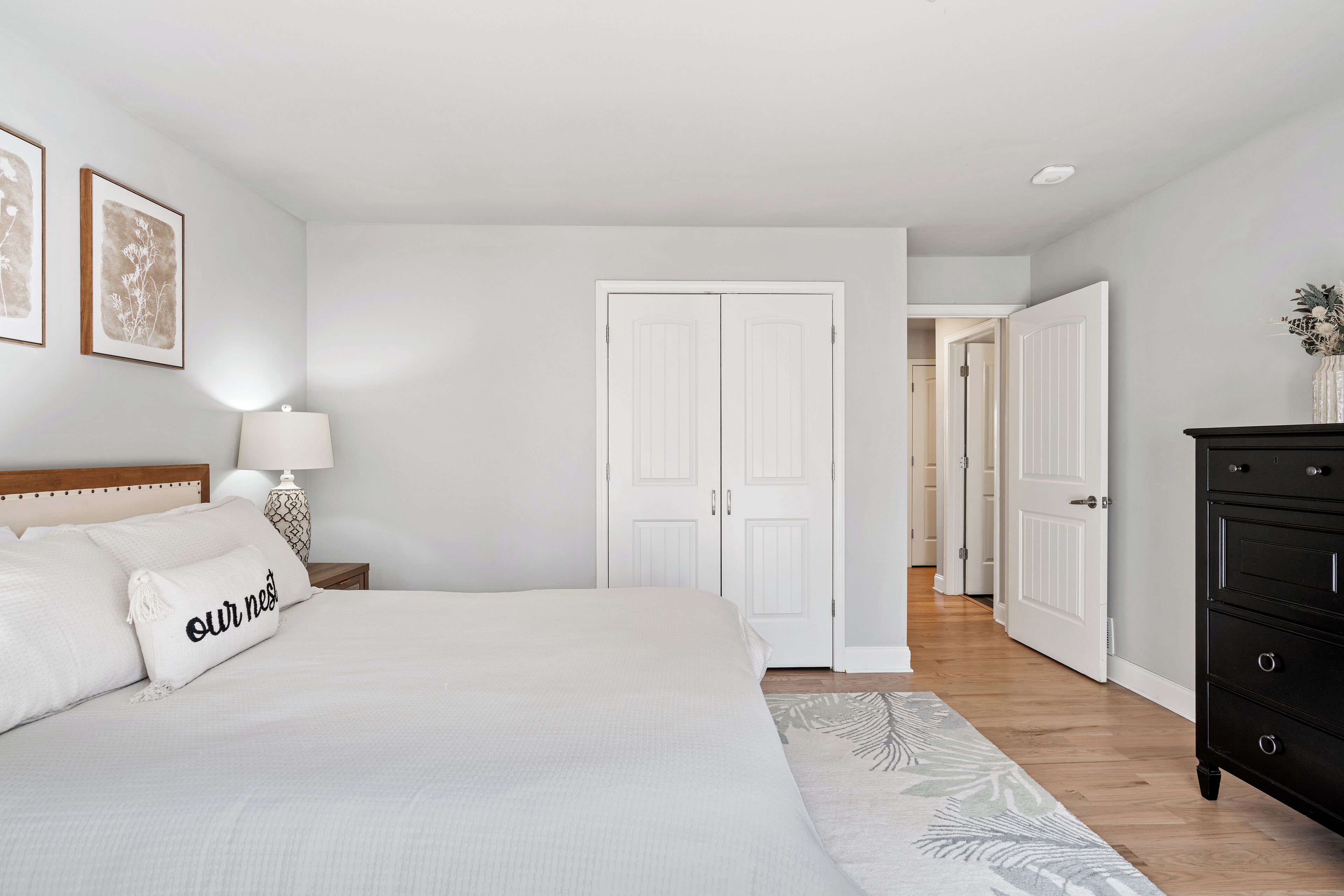 Een slaapkamer met een groot bed, een nachtkastje met een lamp, een dressoir en een deur naar een andere kamer.