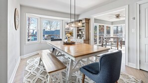 Un espace repas avec une table en bois, des bancs et un lustre. De grandes fenêtres offrent une vue sur l’extérieur.