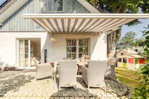 Une terrasse extérieure couverte, aménagée avec des meubles en osier, une table et des chaises, jouxtant un bâtiment doté de grandes fenêtres et d’une porte-fenêtre coulissante.