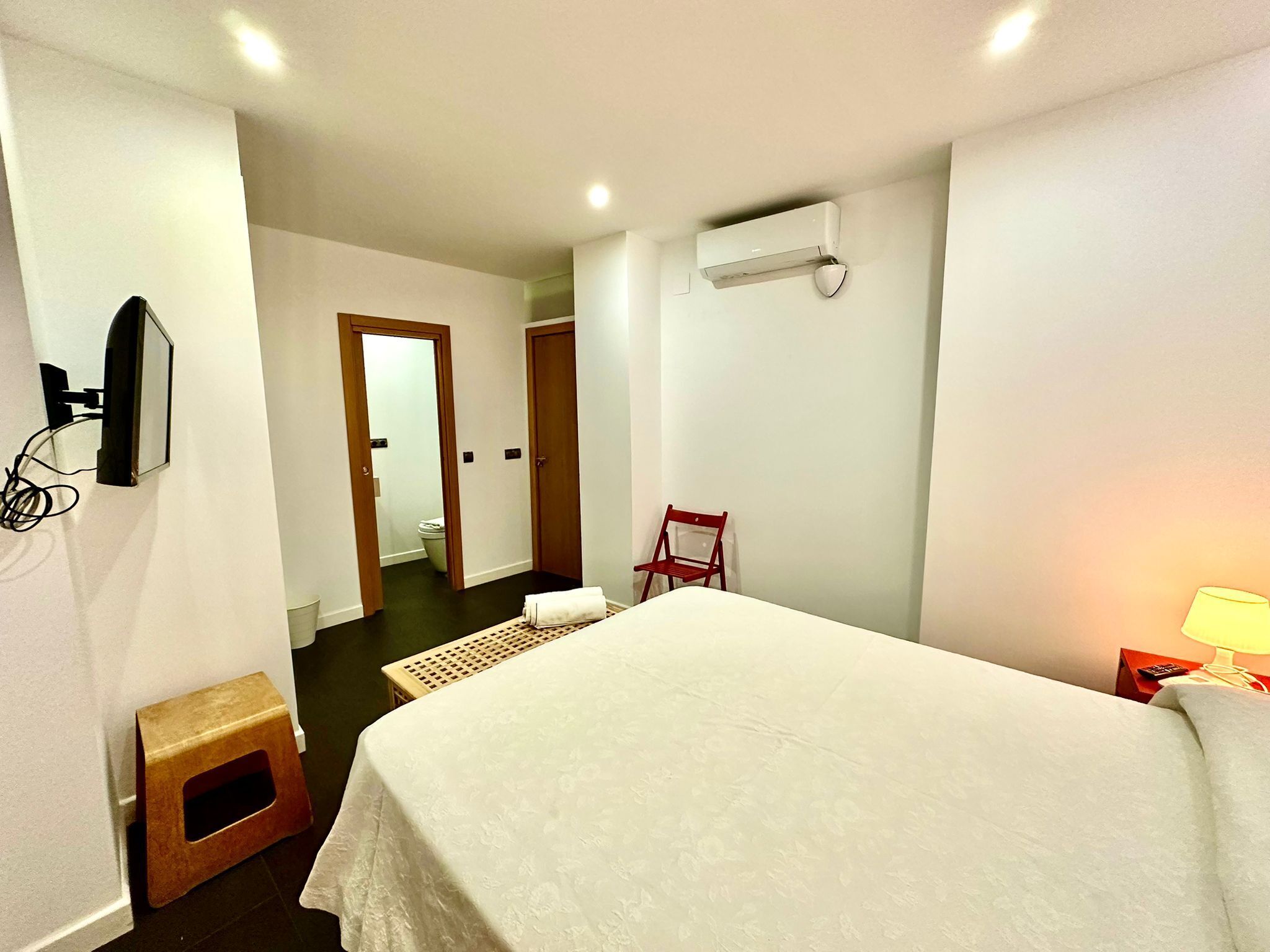 Een hotelkamer met een bed, een klein krukje, een stoel, een aan de muur bevestigde televisie en een airconditioner.