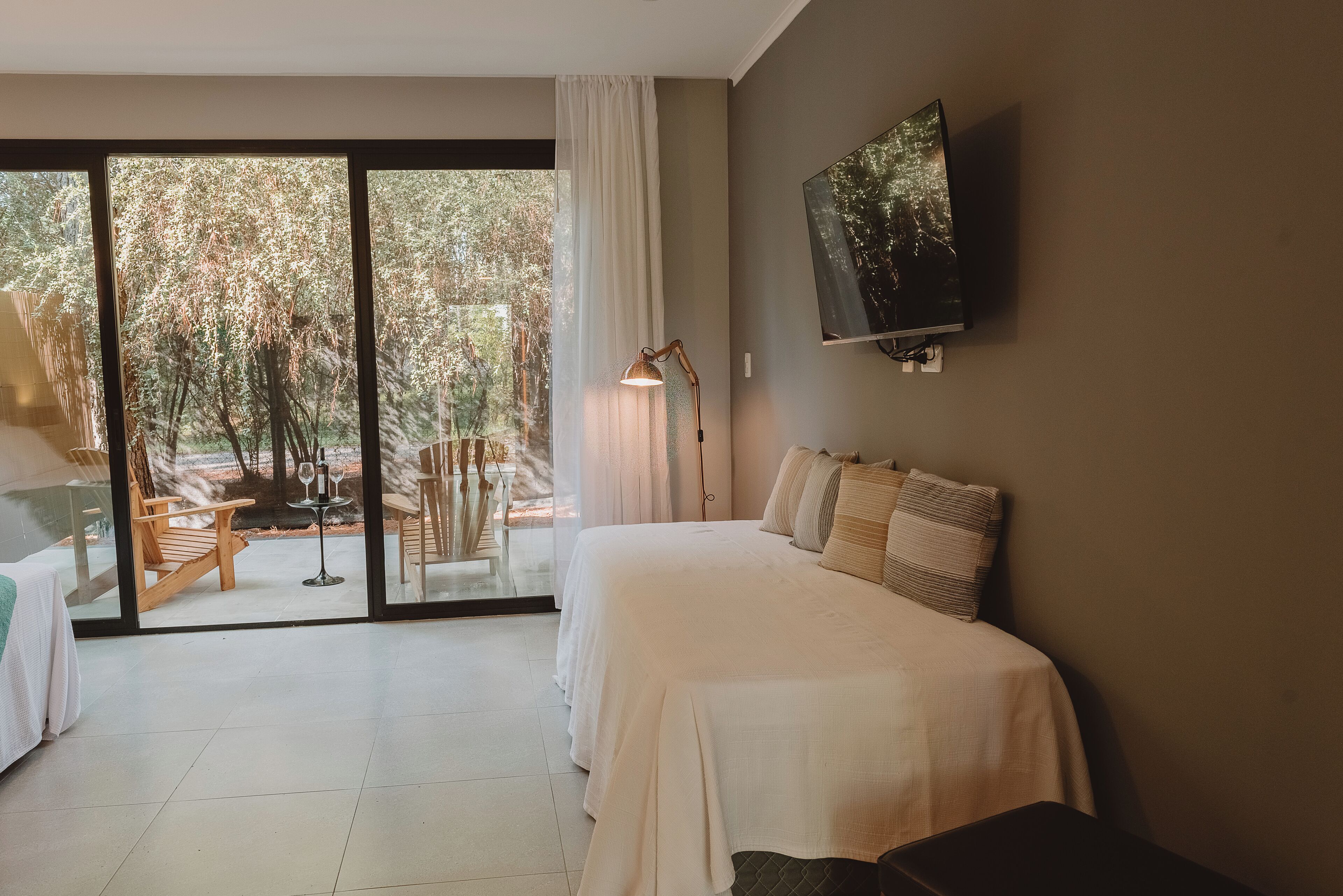 A bedroom with a bed, a TV mounted on the wall, a sliding glass door leading to an outdoor area with a table and chairs, and a lamp on the floor.