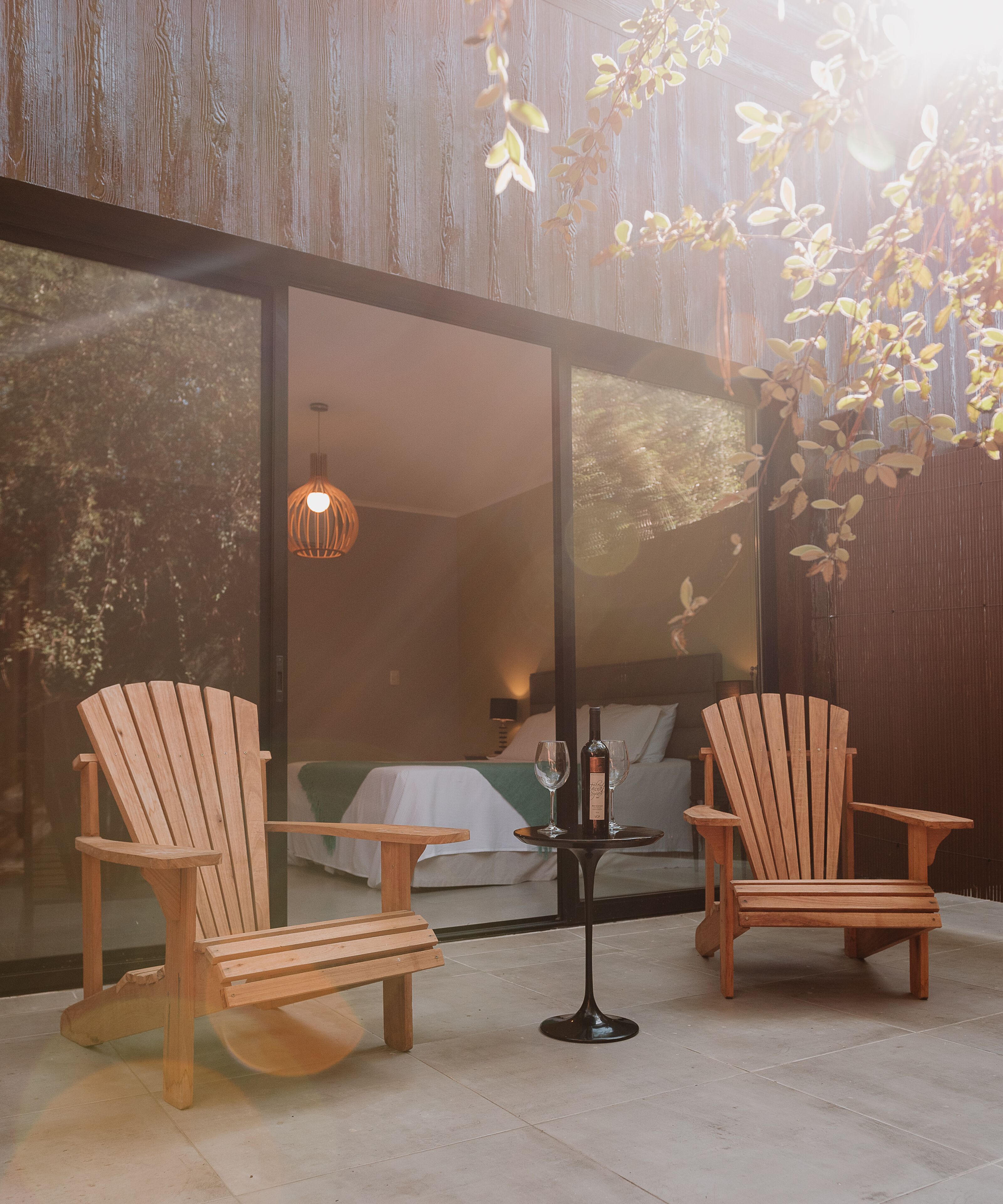 A modern room with two wooden chairs, a small table with a bottle and glasses, and a bed with a green blanket.