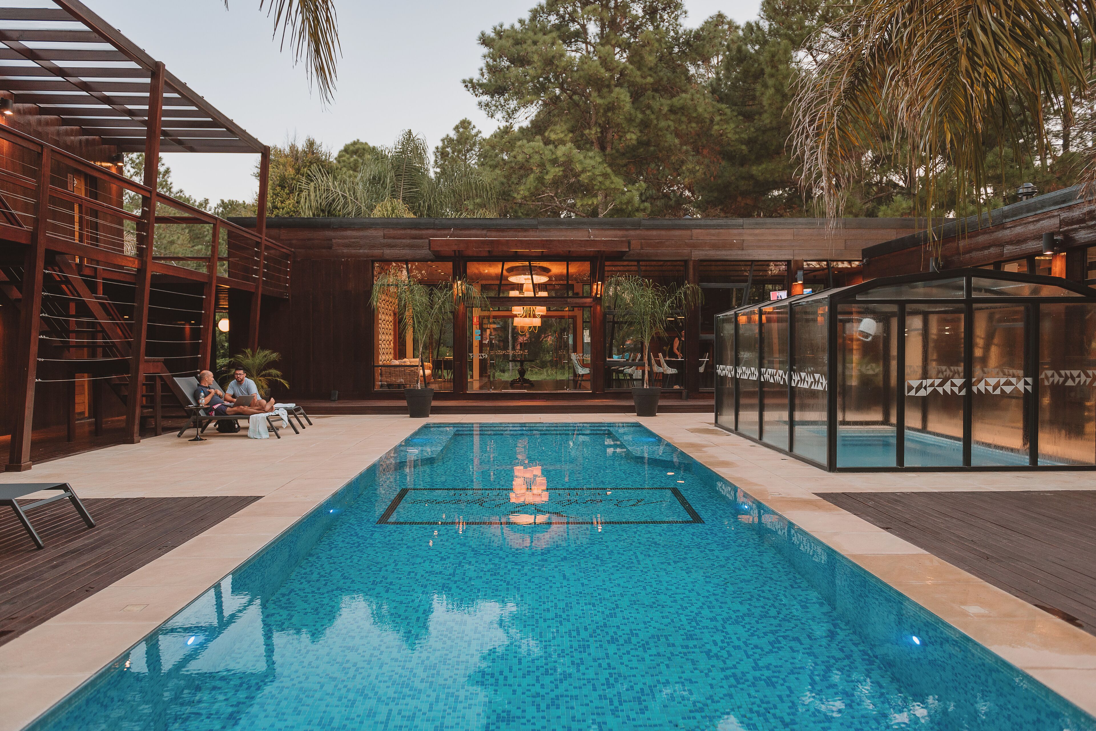 Una piscina con terraza de madera, sillas de descanso y un área de estar cubierta.