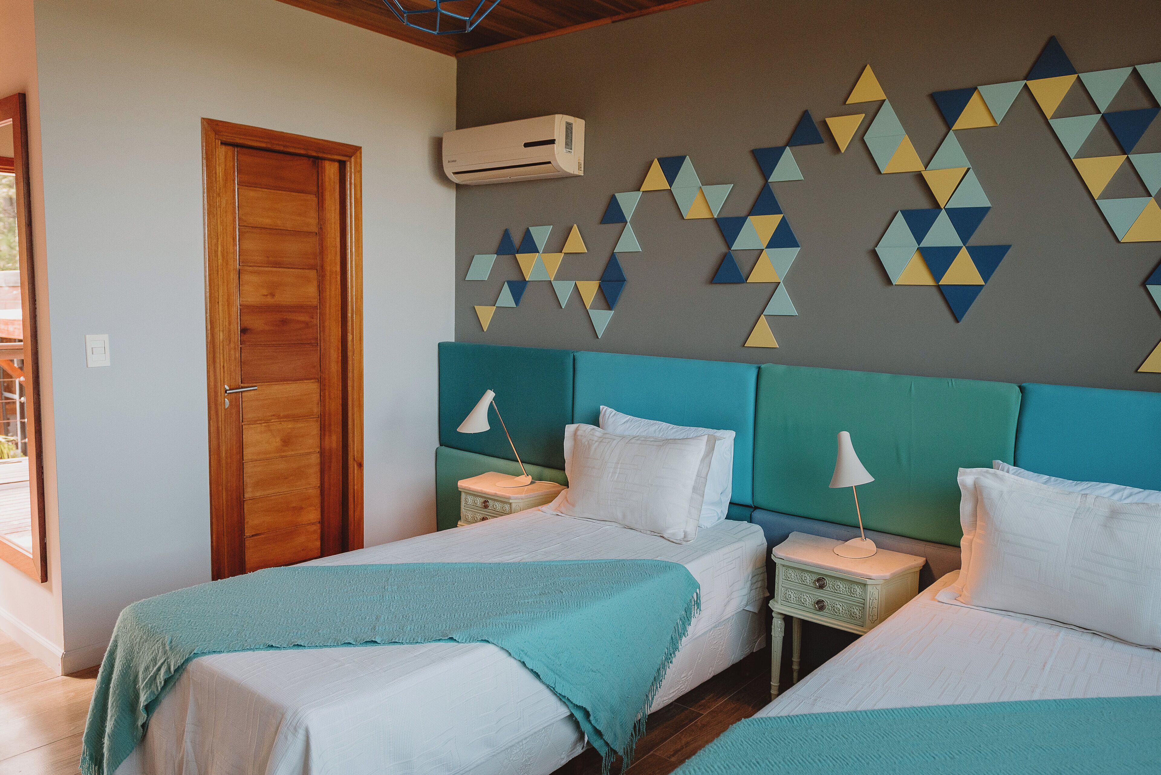 Un dormitorio con dos camas, una puerta de madera, aire acondicionado de pared y decoración mural geométrica.