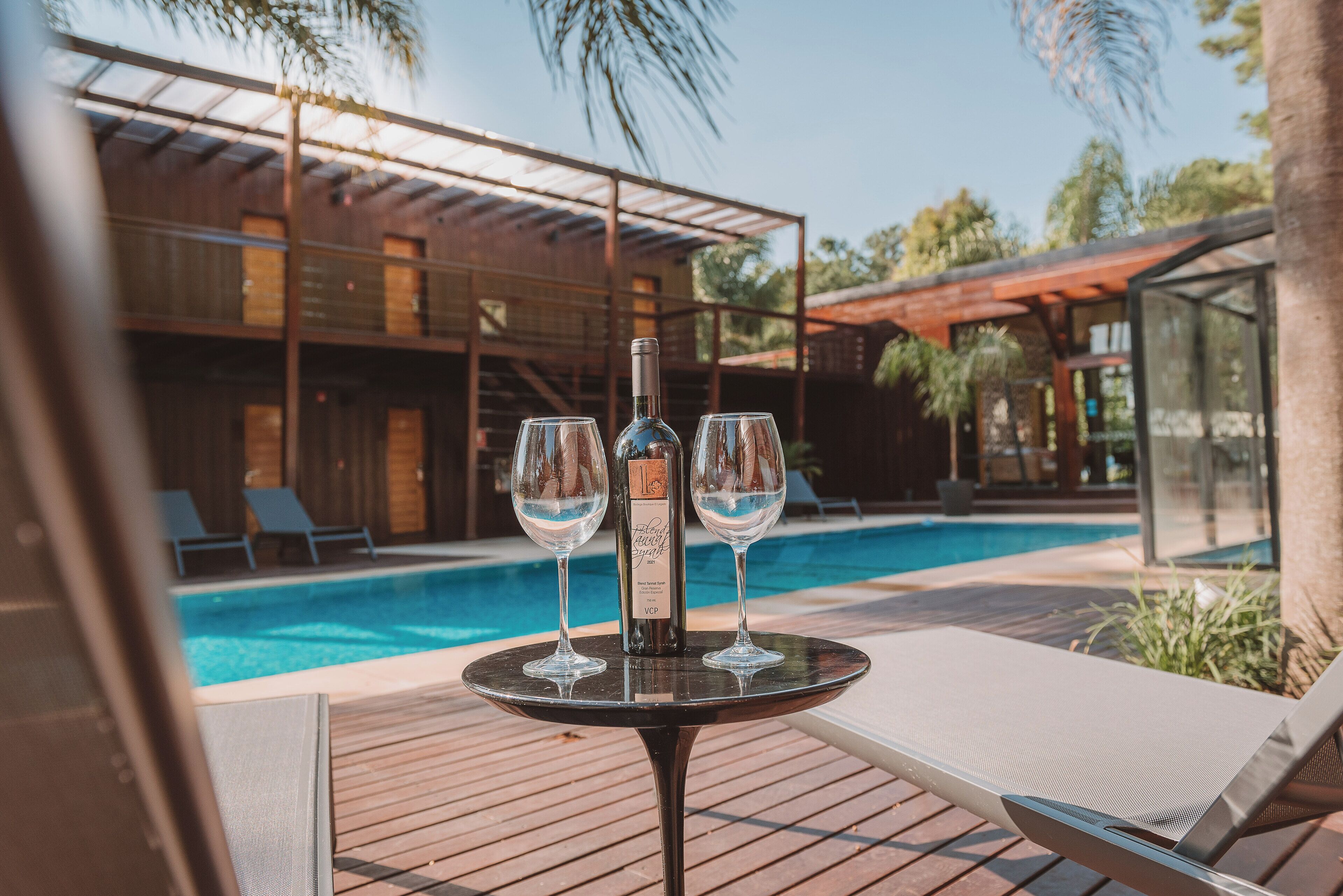 Mesa junto a la piscina con una botella y dos vasos, con vista a una piscina y un edificio moderno con cerramiento de vidrio.