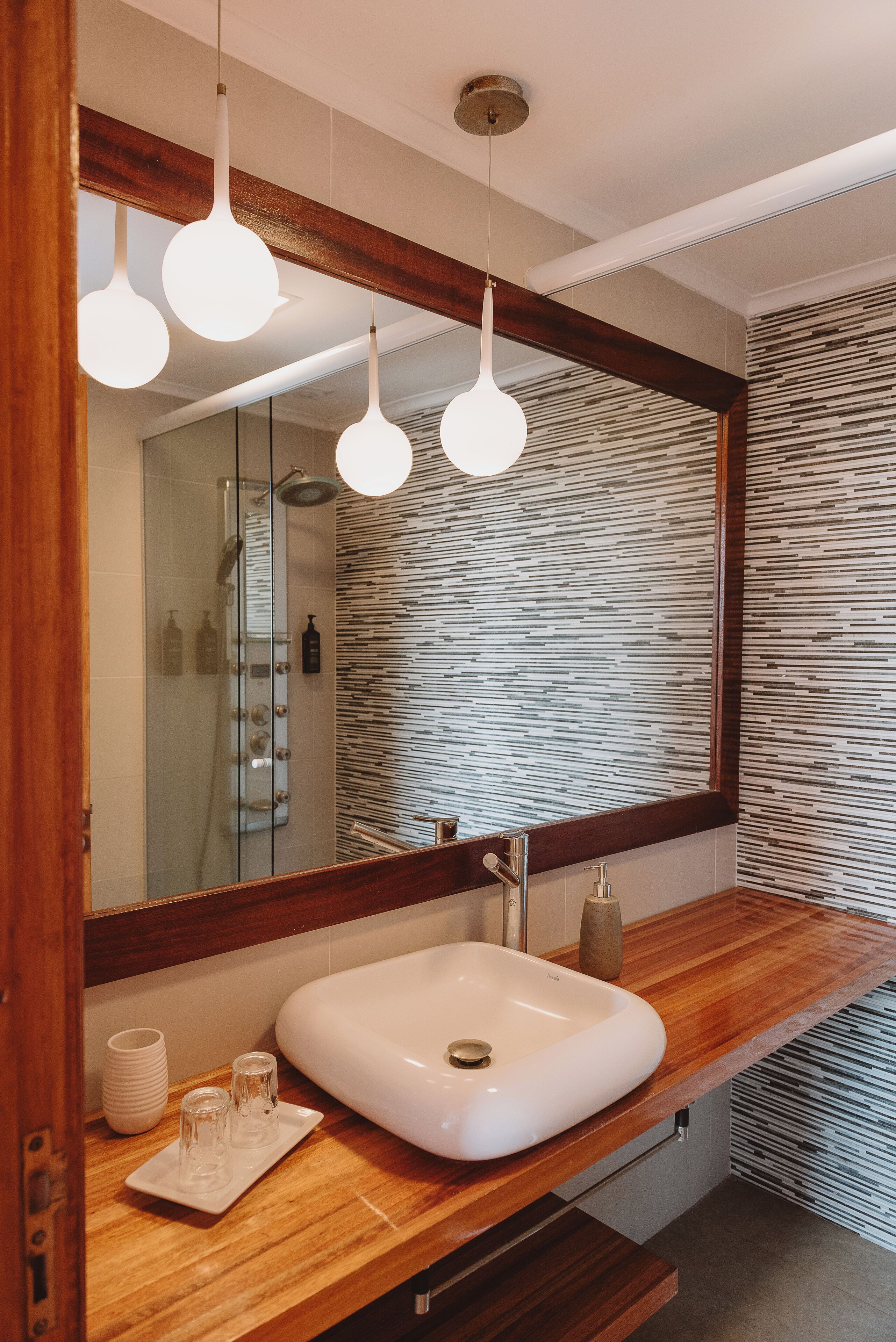 Un baño con tocador de madera, un lavabo blanco y un espejo grande con marco de madera. Hay tres lámparas colgantes sobre el espejo, y una pared texturizada detrás.