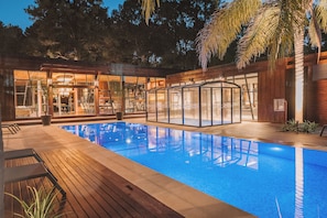 Un área de piscina exterior moderna con cubierta de madera, cerramientos de vidrio y un espacio interior bien iluminado.