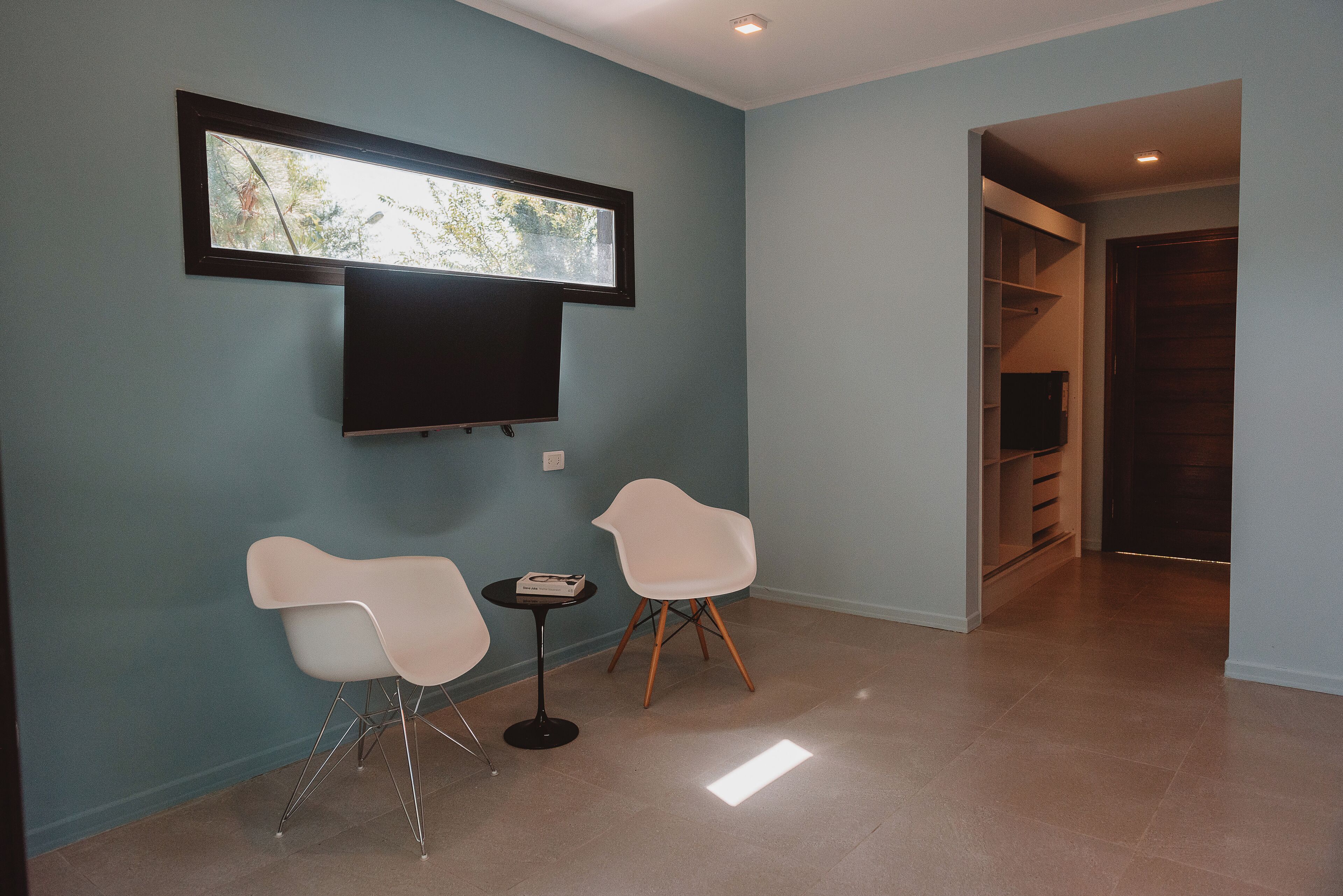 A room with teal walls, a flat-screen TV mounted above a small table with two white chairs, and a view of trees through a window.