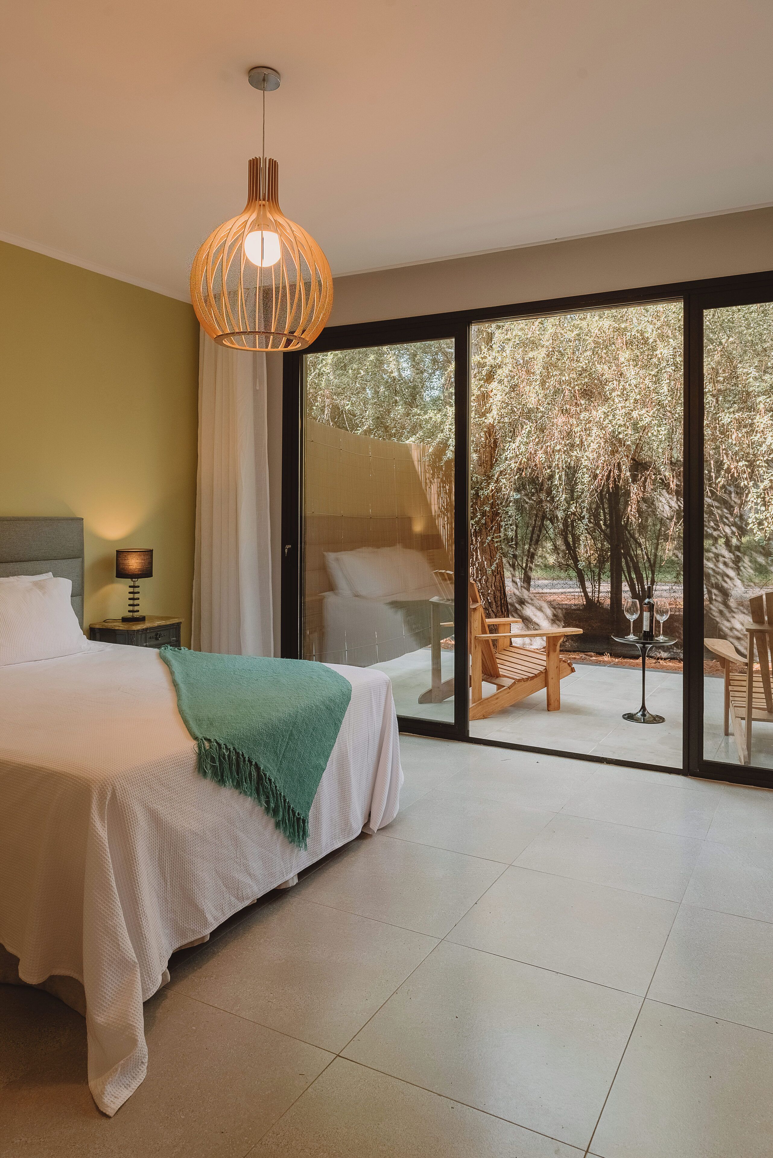 Un dormitorio con cama, mesita de noche con lámpara, manta verde y un amplio ventanal corredero que da a una zona exterior con mesa y sillas.