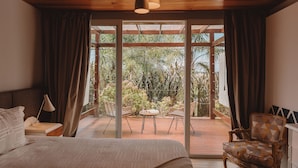 Un dormitorio con cama, mesita de noche, lámpara, silla y vistas a un balcón con plantas y una terraza de madera.