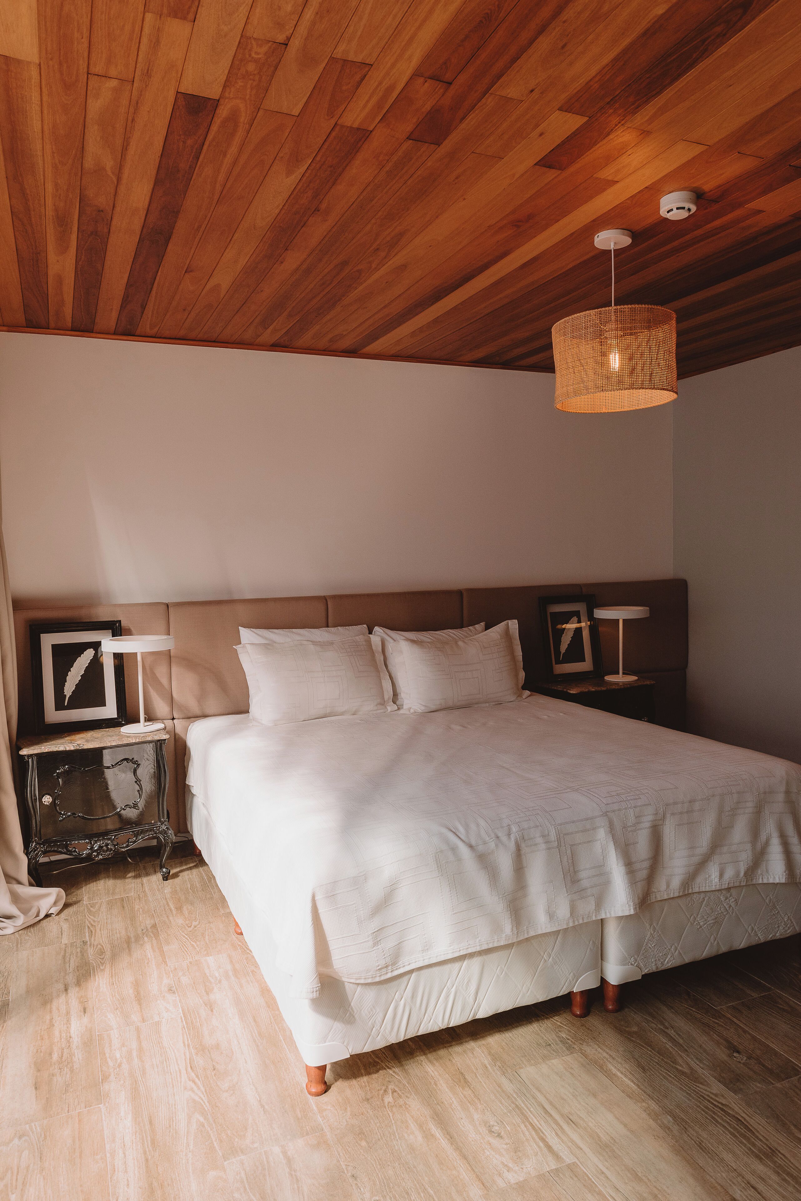 Un dormitorio con techo de madera, una cama con ropa blanca, una mesita de noche con lámpara y un cuadro enmarcado en la pared.