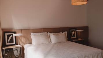 Un dormitorio con techo de madera, una cama con ropa blanca, una mesita de noche con lámpara y un cuadro enmarcado en la pared.