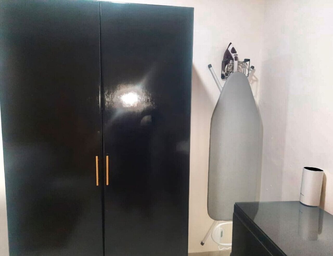 Una habitación con un armario negro, una tabla de planchar gris y una pequeña mesa con un objeto blanco encima.