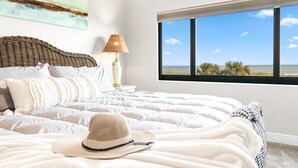A bedroom with a large bed, a view of the ocean through a window, and a painting on the wall. - Wake up to Waves: Stunning Direct Ocean Front View with Heated Pool & Hot Tub! (Cocoa Beach)