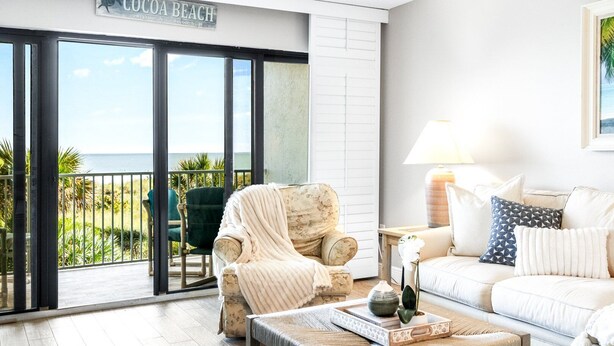 A living room with a view of the ocean, a sofa, a chair, and a coffee table with a decorative item. - Wake up to Waves: Stunning Direct Ocean Front View with Heated Pool & Hot Tub! (Cocoa Beach)