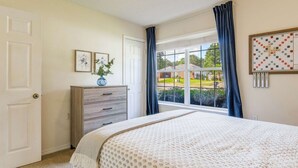 Un dormitorio con cama, cómoda, ventana con cortinas y un crucigrama enmarcado en la pared.