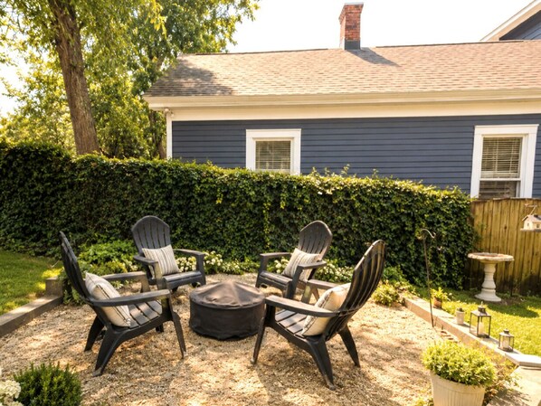 A patio with four Adirondack chairs and a round table, surrounded by a hedge and a wooden fence.