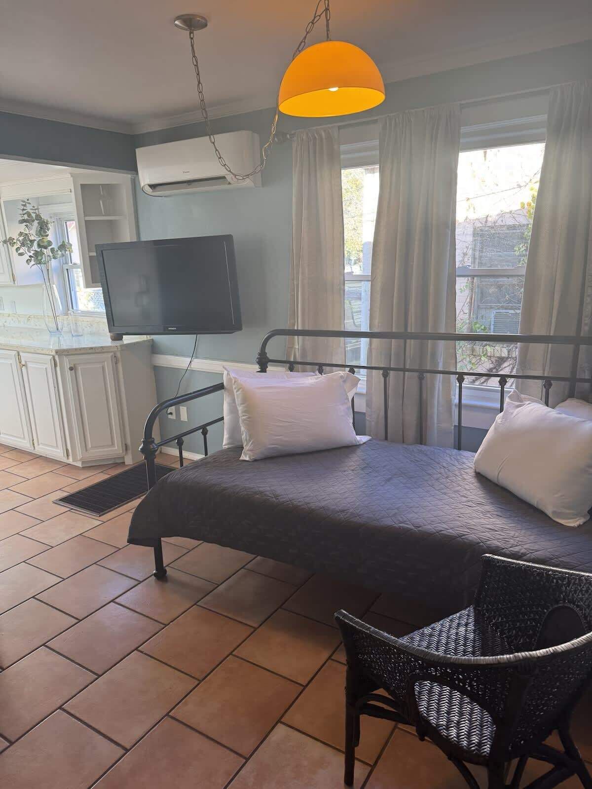 Un dormitorio con una cama, una silla, un televisor y un ventanal con cortinas.