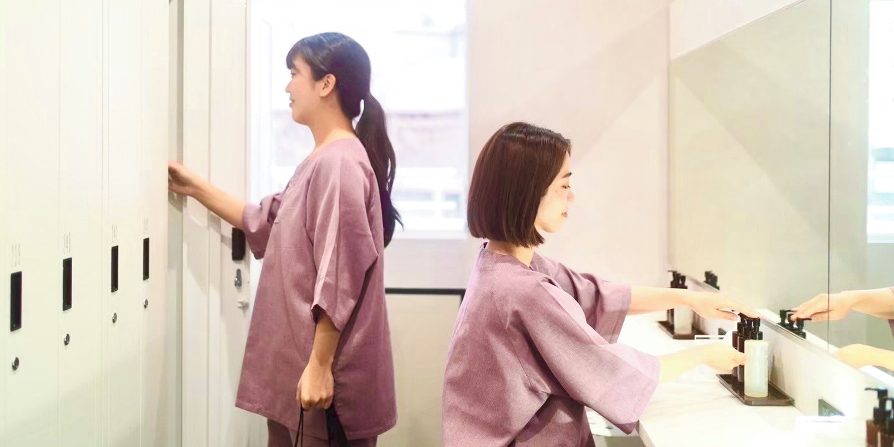 A woman in a purple robe standing in front of a mirror, reaching for a bottle on a counter.