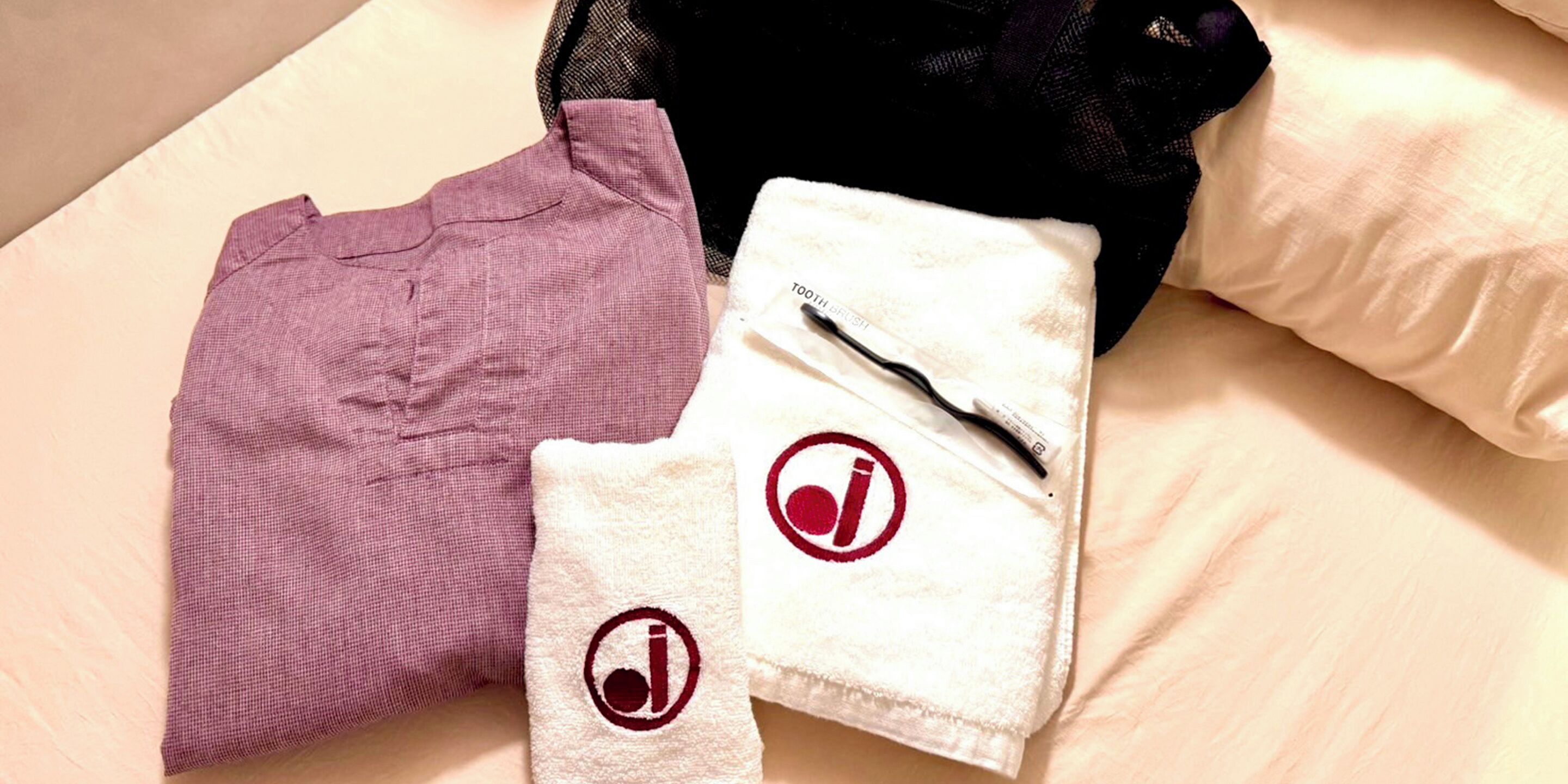 A neatly made bed with a white pillow, a purple garment, and a white towel with a logo.