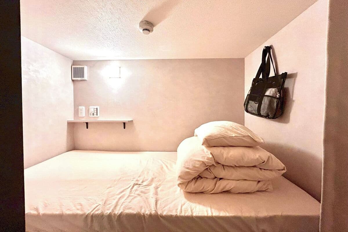 A small, single-bed room with a plain wall, a shelf with a bag, and a ceiling vent.
