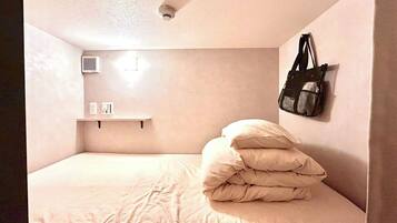 Une petite chambre avec un lit simple, un mur uni, une étagère avec un sac et une bouche d’aération au plafond.