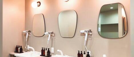 A modern bathroom with three sinks, each with a mirror above. There are hair dryers and a hair straightener on the wall.