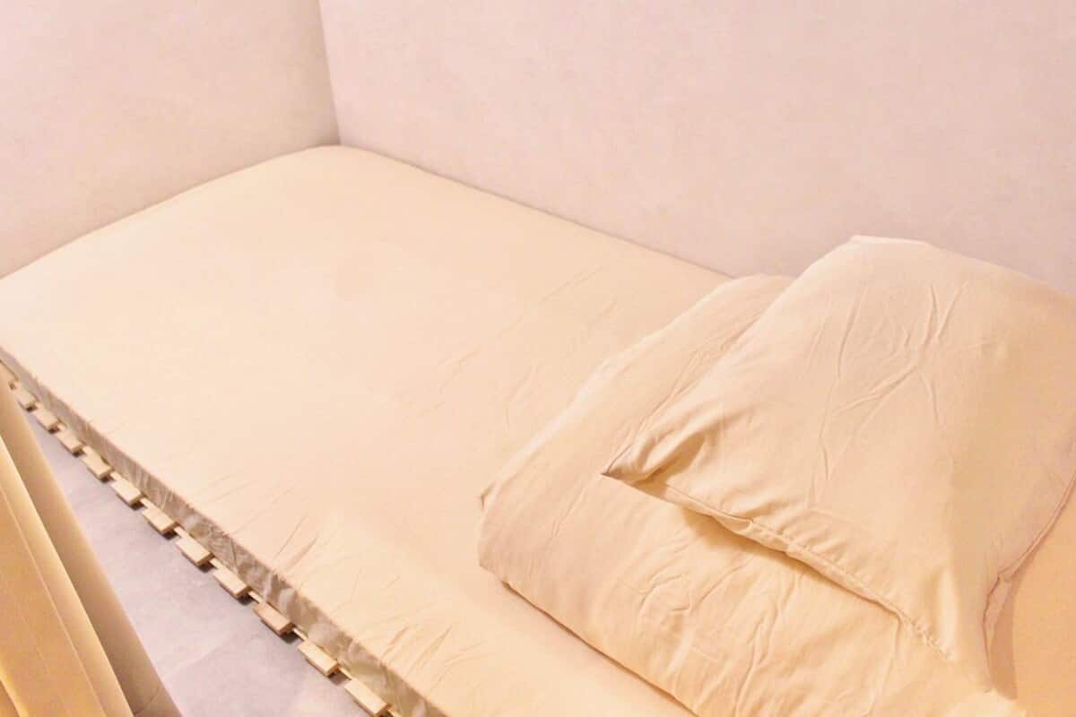A single bed with a beige mattress and pillows, a wooden headboard, and a plain wall.