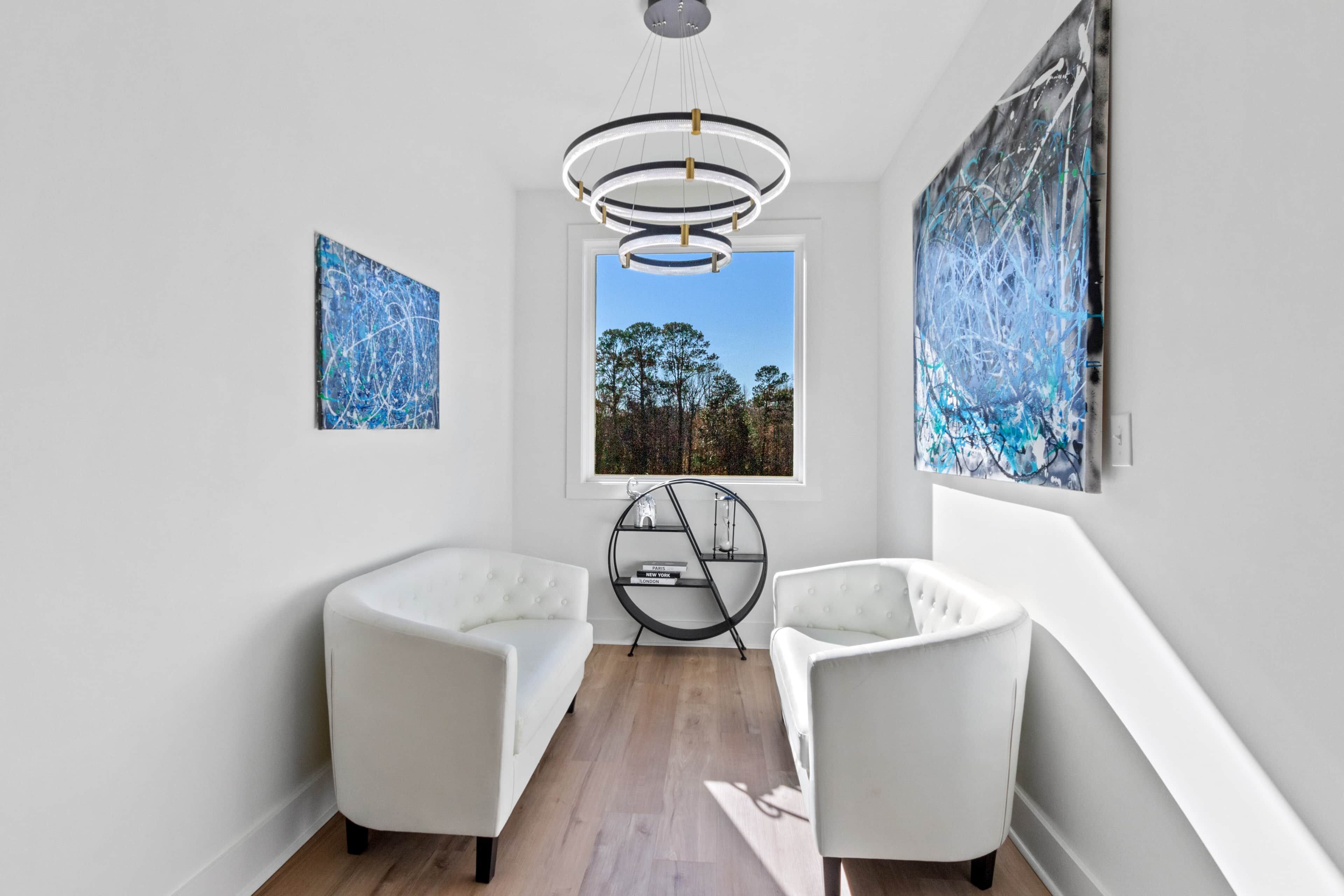 A modern interior with two white armchairs, a round metal shelf, and abstract paintings.