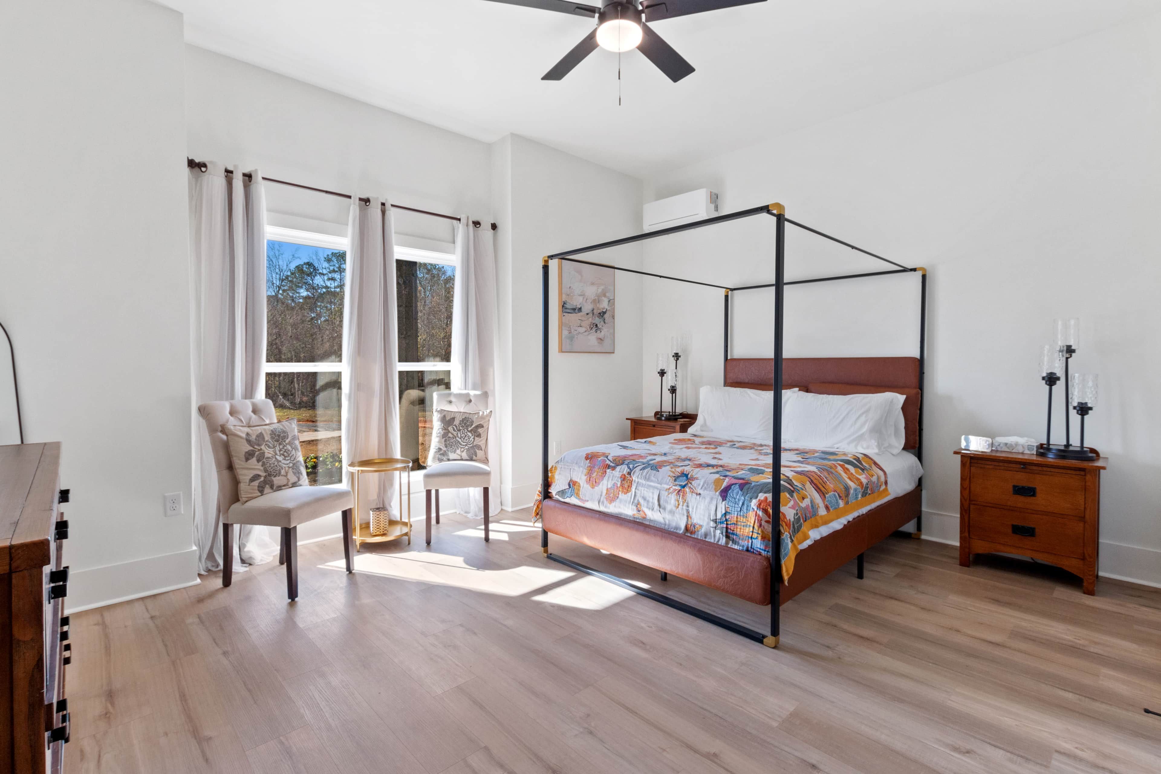 A bedroom with a four-poster bed, a bedside table with a lamp, a chair, and a window with curtains.