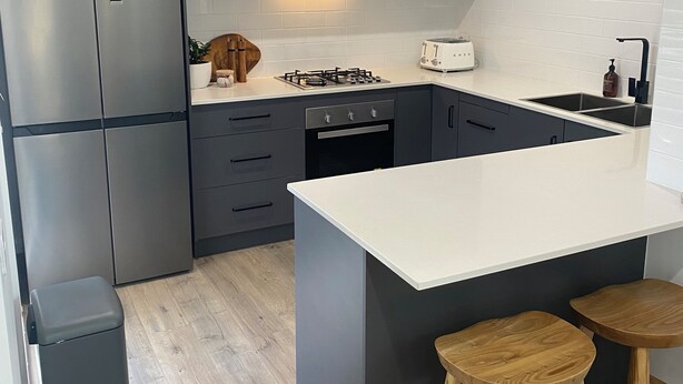 A modern kitchen with grey cabinets, a white countertop, a built-in stove, a sink, and two wooden stools.