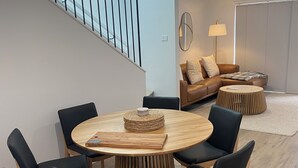 A modern living room with a round wooden dining table surrounded by black chairs, a brown leather sofa, and a small wooden coffee table. A staircase with a railing is visible in the background.