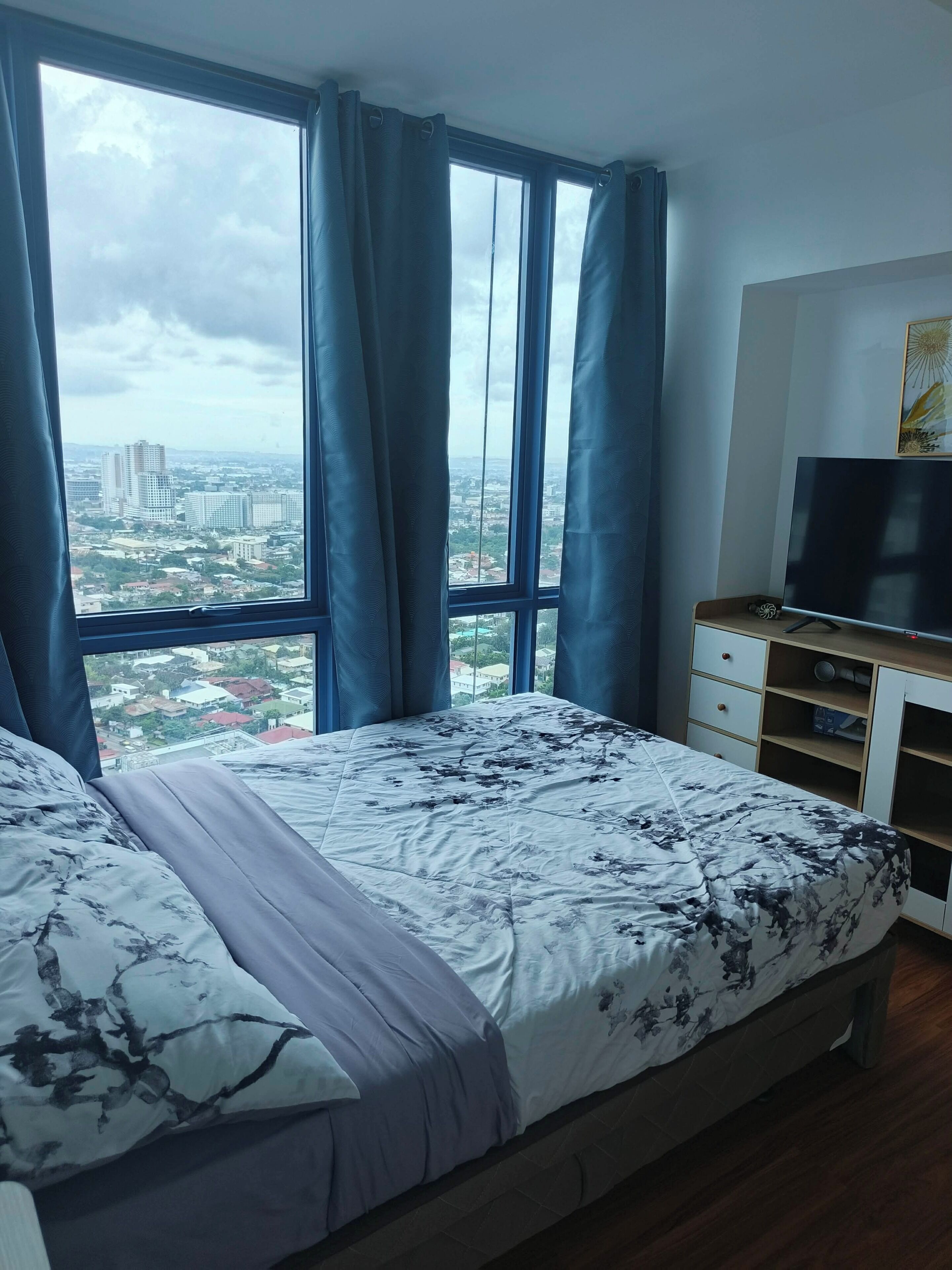 A bedroom with a large bed, a TV, and a view of a cityscape through a window with curtains.