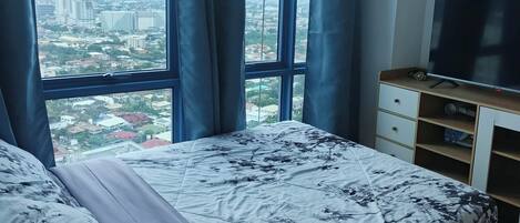 A bedroom with a large bed, a TV, and a view of a cityscape through a window with curtains.