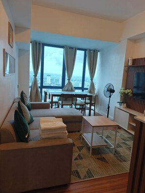 A modern living room with a sofa, a dining table, and chairs. There is a large window with curtains, a flat-screen TV, and a fan.