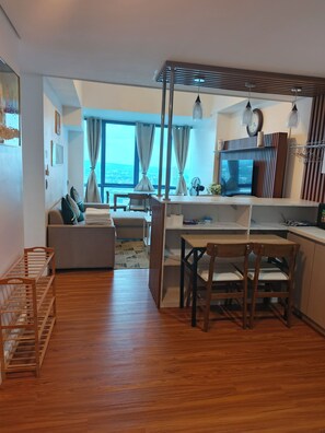 A modern living room with a sofa, dining table, and chairs, a kitchen area with a TV, and a large window with curtains.