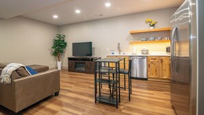 A modern living room with a sofa, TV, and dining area. The kitchen features wooden cabinets, a refrigerator, and a dishwasher. - The E-town Hideaway (Elizabethtown)