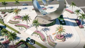 An aerial view of a modern public space with a central sculpture, landscaped areas with palm trees, and a circular pedestrian pathway.